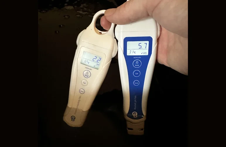 A person holding a digital Bluelab EC meter and pH pen dipped into a dark bucket of cannabis nutrient water, showing readings of 2.2 EC and 5.7 pH.