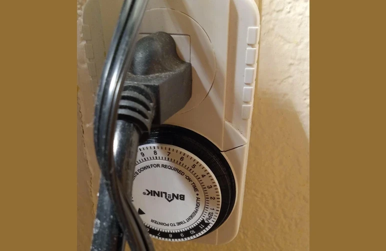 A close up of a white BN-LINK mechanical 24-hour outlet timer plugged into a wall with a black power cord attached, used to automate indoor grow light schedules.